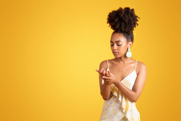 Sad young black woman looks at her hand, need new manicure, suffers from pain and injury, isolated on yellow background, studio. Health problems, nails care, ad and offer, beauty care - Foto, Imagen