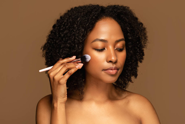 Attractive half-naked young black woman with bushy hair holding makeup brush, isolated on brown background. African american lady applying blush or foundation on her face - Foto, afbeelding