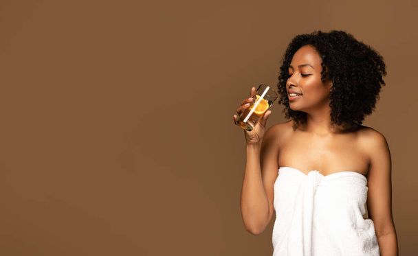 Attraente signora afro-americana millenaria calda avvolta in asciugamano bianco con bicchiere d'acqua con fetta di arancio, isolata su sfondo marrone, panorama con spazio copia. Concetto di idratazione - Foto, immagini