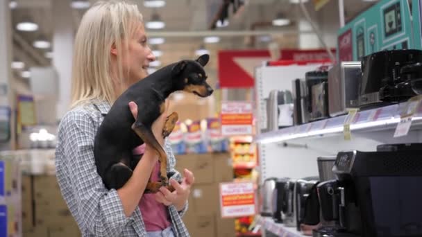 Giovane bella bionda con il suo piccolo cane giocattolo terrier cammina intorno supermercato o negozio e raccoglie elettrodomestici. Famiglia, liberta 'dai bambini. Bambino gratis - Filmati, video