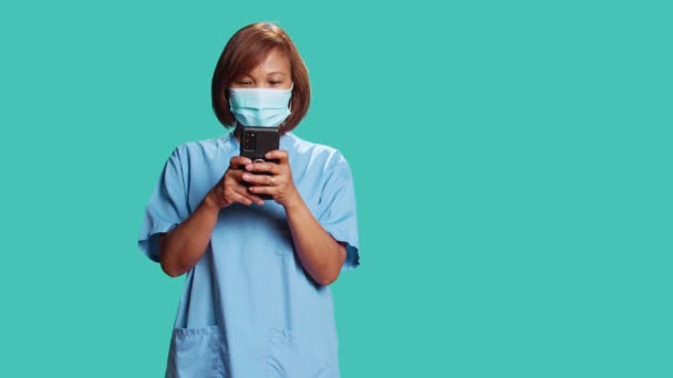 Счастливая расслабленная медсестра, общаясь с друзьями в Интернете во время больничной смены. Улыбающаяся женщина печатает сообщения по телефону, носит медицинскую форму, изолирована на фоне синей студии - Кадры, видео