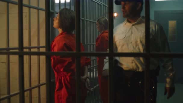Female prisoners, inmates in orange uniforms stand facing the metal bars in front of prison cells. Jailer walks, talks to women, watches criminals in jail. Detention center or correctional facility. - Felvétel, videó