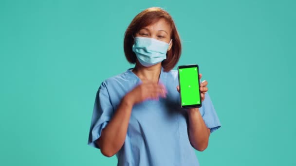 Védőmaszkot viselő nővér orvosi utasításokat mutat videó a telefon zöld képernyőjén. Kórházi alkalmazott gazdaság chroma kulcs mock up okostelefon, elszigetelt felett kék stúdió háttér - Felvétel, videó