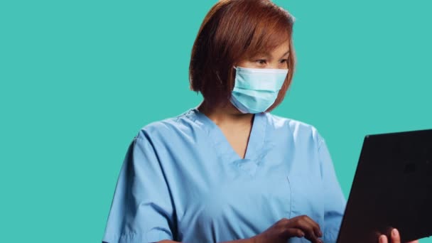 Közelkép mosolygós boldog egészségügyi szakember segítségével laptop keresni orvosi információk online. Ázsiai nővér visel professzionális klinikai védő egyenruhát, izolált stúdió háttér - Felvétel, videó