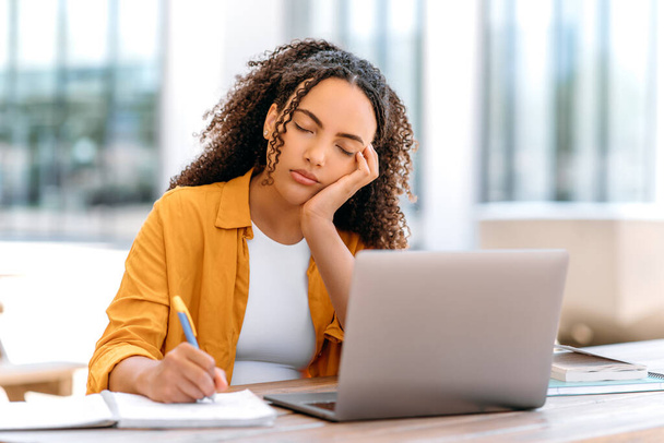 Müde junge Frau mit lockigem Haar, gemischte Rasse, sitzt draußen mit Laptop, schläft ein, während sie studiert oder online arbeitet, fühlt sich müde und schläft nicht, schläft mit geschlossenen Augen - Foto, Bild
