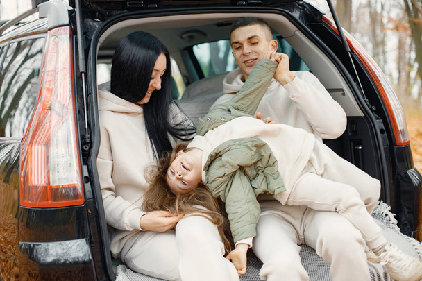 Family resting after day spending outdoor in autumn park. Father, mother, their daughter hugging inside car trunk, smiling. Family wearing beige sportive costumes. - Photo, image