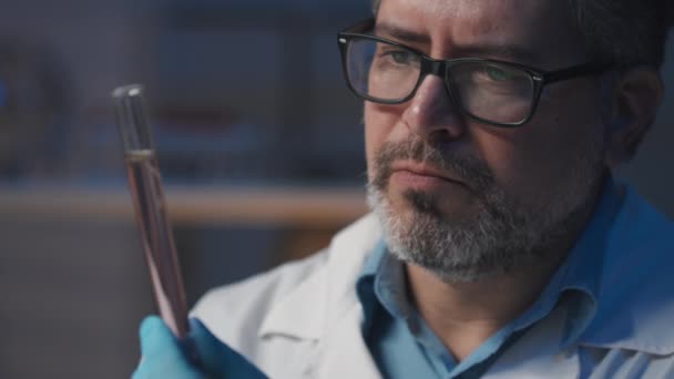 Primer plano del ingeniero genético masculino serio en anteojos mirando frasco con plántulas líquidas y diminutas de plantas cultivadas durante el experimento científico en laboratorio - Imágenes, Vídeo