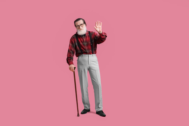 Senior man with walking cane waving on pink background - Φωτογραφία, εικόνα