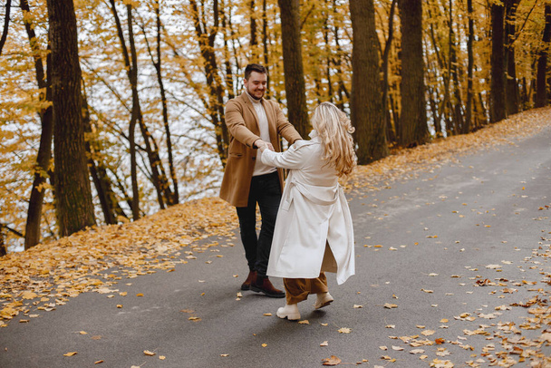 Young man and woman walking outside wearing beige coats. Blond woman and brunette man in autumn forest. Romantic couple holding hands. - Foto, immagini