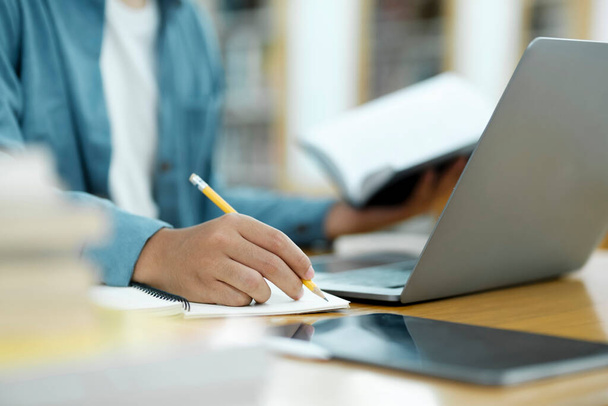Üniversite Kütüphanesi: Zeki Asyalı üniversite öğrencisi Laptop kullanır, Kağıt için Notlar Yazar, Sınıf ödevi için Çalışma. Odaklanmış öğrenciler öğreniyor, üniversite sınavlarına çalışıyor.. - Fotoğraf, Görsel