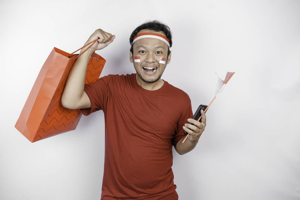 An attractive Asian man standing excited holding an online shopping bag and his smartphone, studio shot isolated on white background. Indonesia's Independence day concept - Foto, imagen
