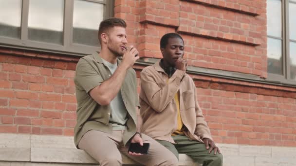 Slowmo of two diverse male friends smoking electronic devices and having conversation outdoors - Materiał filmowy, wideo