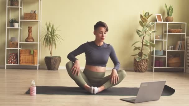 Contemporary young pregnant woman rolling from one side to another on mat while having online sports training via laptop - Imágenes, Vídeo