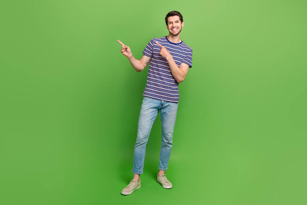 Full length photo of positive funky guy wear striped t-shirt pointing two fingers empty space isolated green color background. - Φωτογραφία, εικόνα