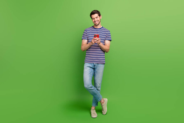 Full length photo of positive cute guy wear striped t-shirt communicating modern gadget isolated green color background. - Foto, immagini