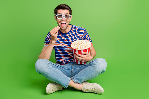 Full length photo of impressed excited guy wear striped t-shirt watching film eating pop corn empty space isolated green color background. - Photo, Image