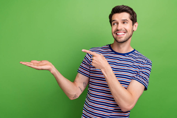 Photo of charming cheerful man dressed striped t-shirt pointing finger holding arm empty space isolated green color background. - Valokuva, kuva