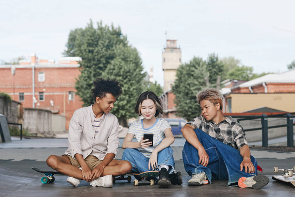 Retrato de comprimento total de grupo diversificado de três inquilinos no parque de skate ao ar livre sentado no chão e usando o ambiente urbano do smartphone - Foto, Imagem