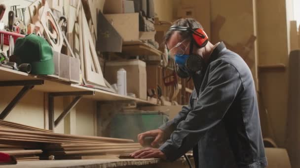 Μεσαία βολή του επαγγελματία ξυλουργού σε μάσκα ασφαλείας γυάλισμα ξύλινες σανίδες στο γραφείο στο εργαστήριο - Πλάνα, βίντεο