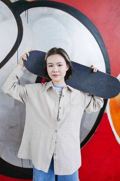 Κάθετη πορτρέτο της νεαρής έφηβης με skateboard στέκεται στον τοίχο γκράφιτι στο κόκκινο και κοιτάζοντας την κάμερα - Φωτογραφία, εικόνα