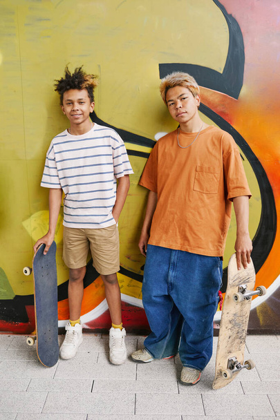 緑の落書きの壁に立って,カメラを見ているスケートボードを持つ2人のティーンエイジャーの男の子の垂直の肖像画 - 写真・画像