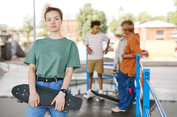Tailliertes Porträt eines Teenager-Mädchens mit Skateboard, das in die Kamera blickt, diverse Freundesgruppen im Hintergrund, Kopierraum - Foto, Bild