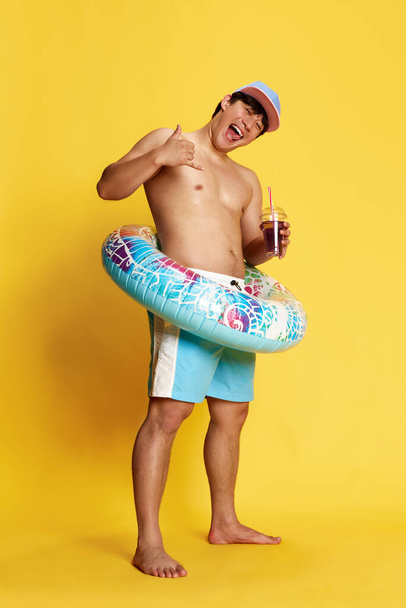 Full-length image of young man in swimming trunks, standing into swimming circle and drinking cocktail over yellow studio background. Concept of summer, vacation, leisure time, holidays, emotions, ad - Фото, зображення