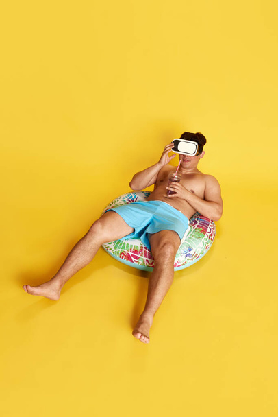 Full-length image of young man in swimming trunks and vr glasses, sitting into swimming circle against yellow studio background. Concept of summer, vacation, leisure time, holidays, human emotions, ad - Photo, Image