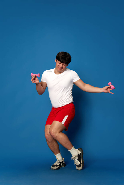 Plný portrét ošuntělého mladého muže v tréninku sportovního oblečení s činkami a baví se na modrém pozadí studia. Pojetí sportu, fitness, aktivního a zdravého životního stylu, lidských emocí, reklam - Fotografie, Obrázek