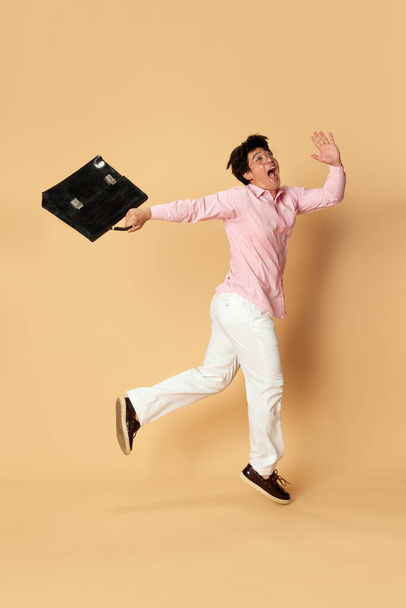 Full-length portrait young man in smart casual clothes jumping with briefcase against yellow studio background. Concept of business, employment, human emotions, office, fashion, ad - Φωτογραφία, εικόνα