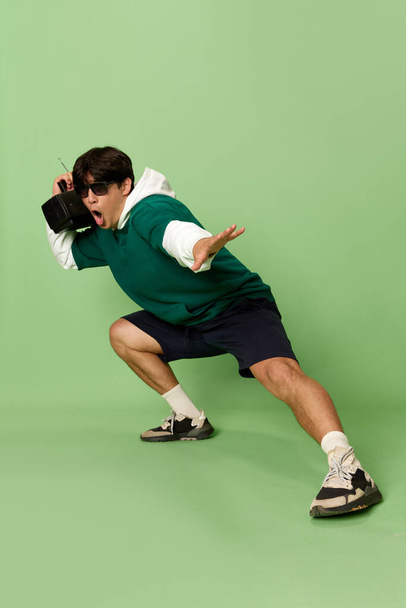 Πορτρέτο του νεαρού κομψό άνδρα σε γυαλιά ηλίου και αθλητικά ρούχα ακούγοντας μουσική σε ρετρό παίκτη πάνω από το πράσινο φόντο στούντιο. Έννοια των ανθρώπινων συναισθημάτων, lifestyle, 90s, μόδα, χαρά, νεολαία, διαφήμιση - Φωτογραφία, εικόνα