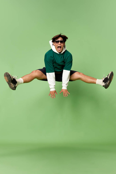 Portrait of young stylish man in sunglasses and sport style clothes cheerfully jumping against green studio background. Concept of human emotions, lifestyle, 90s, fashion, joy, youth, ad - Foto, Bild