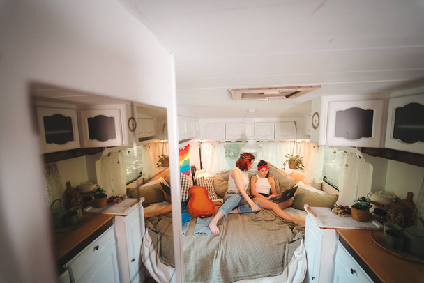 Portrait of a cute lesbian couple. Two girls spend time tenderly together watching movie on laptop in a camper trailer with LGBT flag on the wall. Love and attitude. LGBT concept. High quality photo - Foto, Imagen