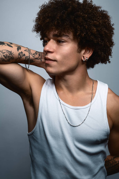 Pensive muscular Brazilian male outlaw gang with curly hair and tattoos with necklace and earrings looking away and smiling against gray background - Fotó, kép