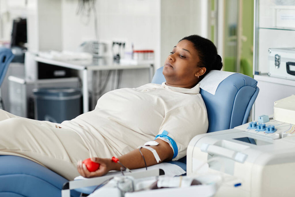 Porträt einer jungen Afroamerikanerin, die Blut spendet, während sie im Stuhl im Plasma-Spendezentrum liegt - Foto, Bild