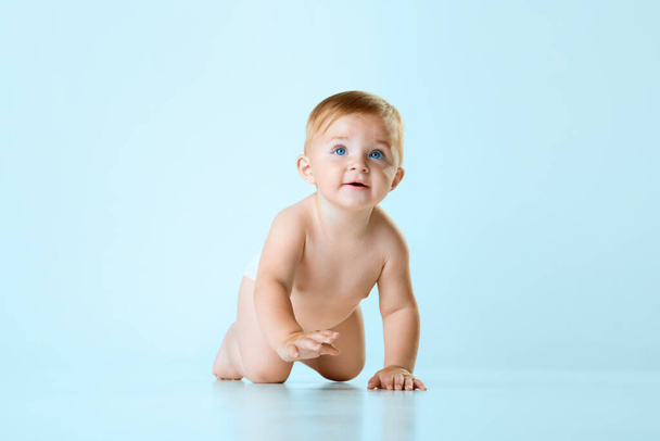 Little cute baby, child, toddler in diaper crawling on floor against light blue studio background. Blue eyes. Concept of childhood, newborn lifestyle, happiness, care. Copy space for ad - Фото, изображение