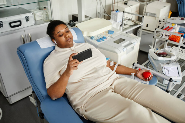 Retrato de alto ángulo de la mujer negra donando sangre mientras está sentada en la silla en el centro de donación de sangre y sosteniendo el teléfono inteligente, espacio para copiar - Foto, imagen