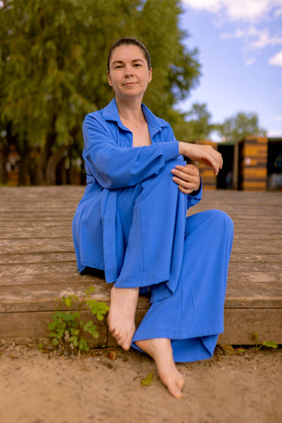 relaxed woman in stylish blue cotton suit sitting on wooden embankment. wrinkle-free travel clothes content. Eco Friendly 100% Cotton Soft Double Layer Crinkle Gauze Muslin Crepe Fabric - Valokuva, kuva