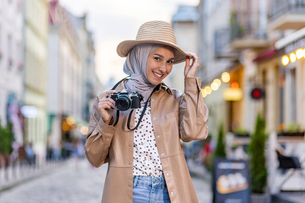 Portrait of young beautiful Muslim woman in hijab and hat, tourist walking in evening city with camera, woman traveling to different countries. - Foto, Bild