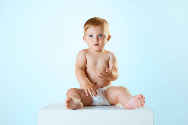 Portret van kleine mooie baby, peuter in luier rustig zitten en kijken tegen lichtblauwe studio achtergrond. Concept van de kindertijd, pasgeboren levensstijl, geluk, zorg. Kopieer ruimte voor advertentie - Foto, afbeelding