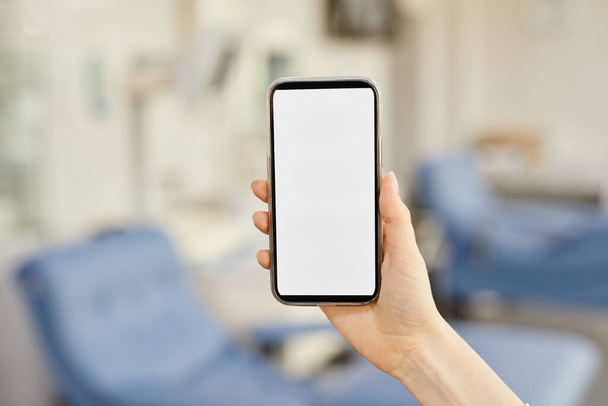 Nahaufnahme einer weiblichen Hand, die ein Smartphone mit leerem Bildschirm im Blutspendezentrum hält, Kopierplatz - Foto, Bild