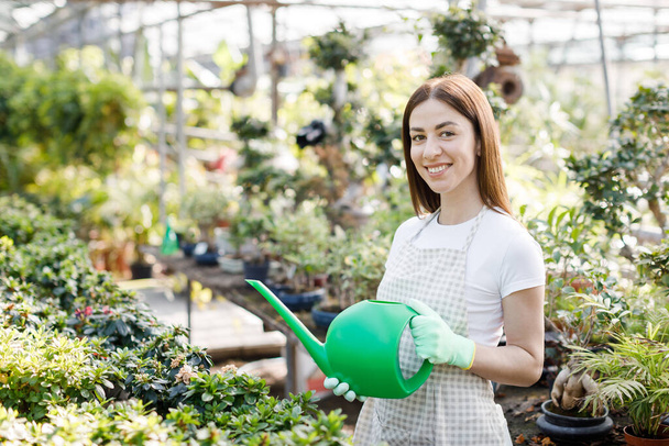 A young woman stands in the middle of a large greenhouse holding a watering can and smiling at the camera - Zdjęcie, obraz