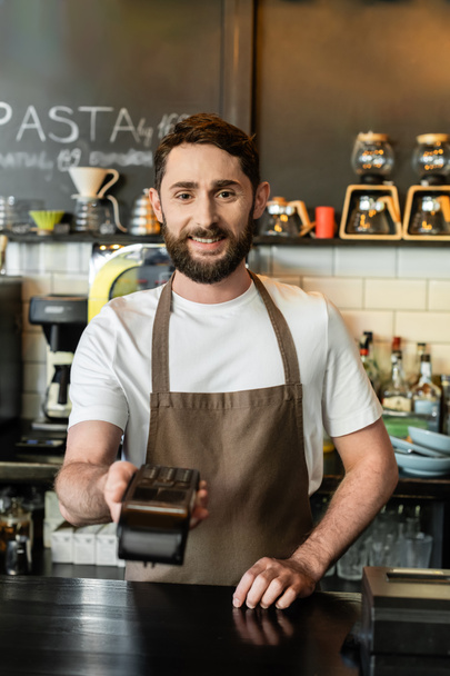 smiling barista in apron holding payment terminal and looking at camera in coffee shop - Photo, Image