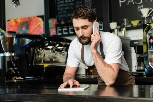 barista en delantal hablando en el teléfono inteligente y el uso de la tableta digital en el bar en la cafetería - Foto, Imagen