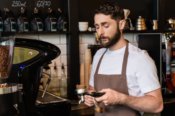 barista barbudo en delantal sosteniendo cepillo y soporte de la máquina de café mientras trabaja en la cafetería - Foto, Imagen