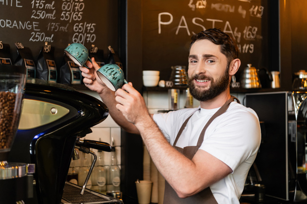 θετική γενειοφόρος barista σε ποδιά κοιτάζοντας κάμερα και κρατώντας κύπελλα κοντά σε καφετιέρα στο καφέ - Φωτογραφία, εικόνα