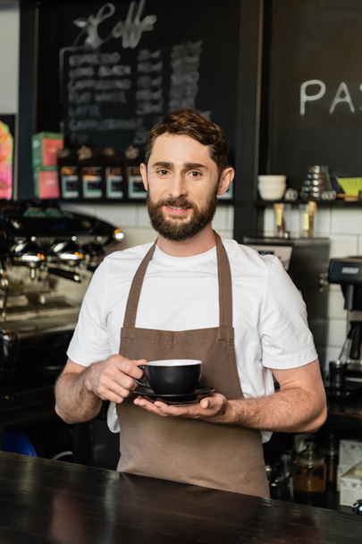 sonriente barbudo barista en delantal sosteniendo taza de café y mirando a la cámara en la cafetería - Foto, Imagen