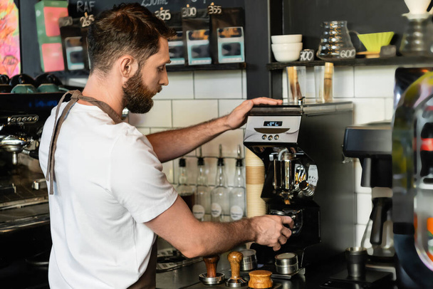 πλαϊνή άποψη του barista σε ποδιά ρίχνει καφέ σε κάτοχο κοντά σε καφετιέρα, ενώ εργάζονται σε cafe - Φωτογραφία, εικόνα
