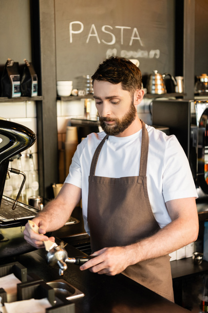 szakállas barista köténytisztító tartó kefével kávéfőző mellett, miközben dolgozik a kávézó - Fotó, kép