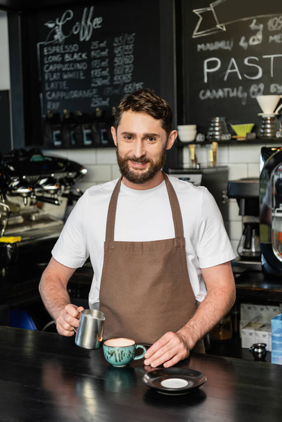 smiling barista in apron holding pitcher with milk near cappuccino in cup in coffee shop - Foto, Imagem